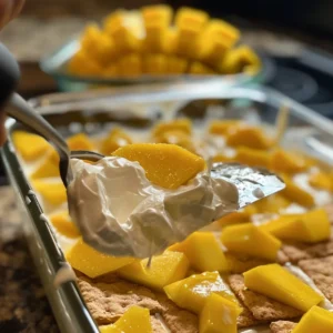 Assembling mango float by layering cream over graham crackers
